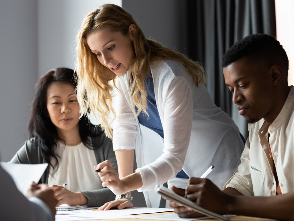 Tre personer vid skrivbord, tittar på papper på bordet. en står lutad över och pratar med de andra